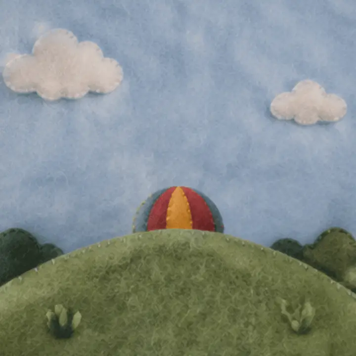 A small felt hot-air balloon rises from behind a green felt hill into a pale felt sky and drifts back down